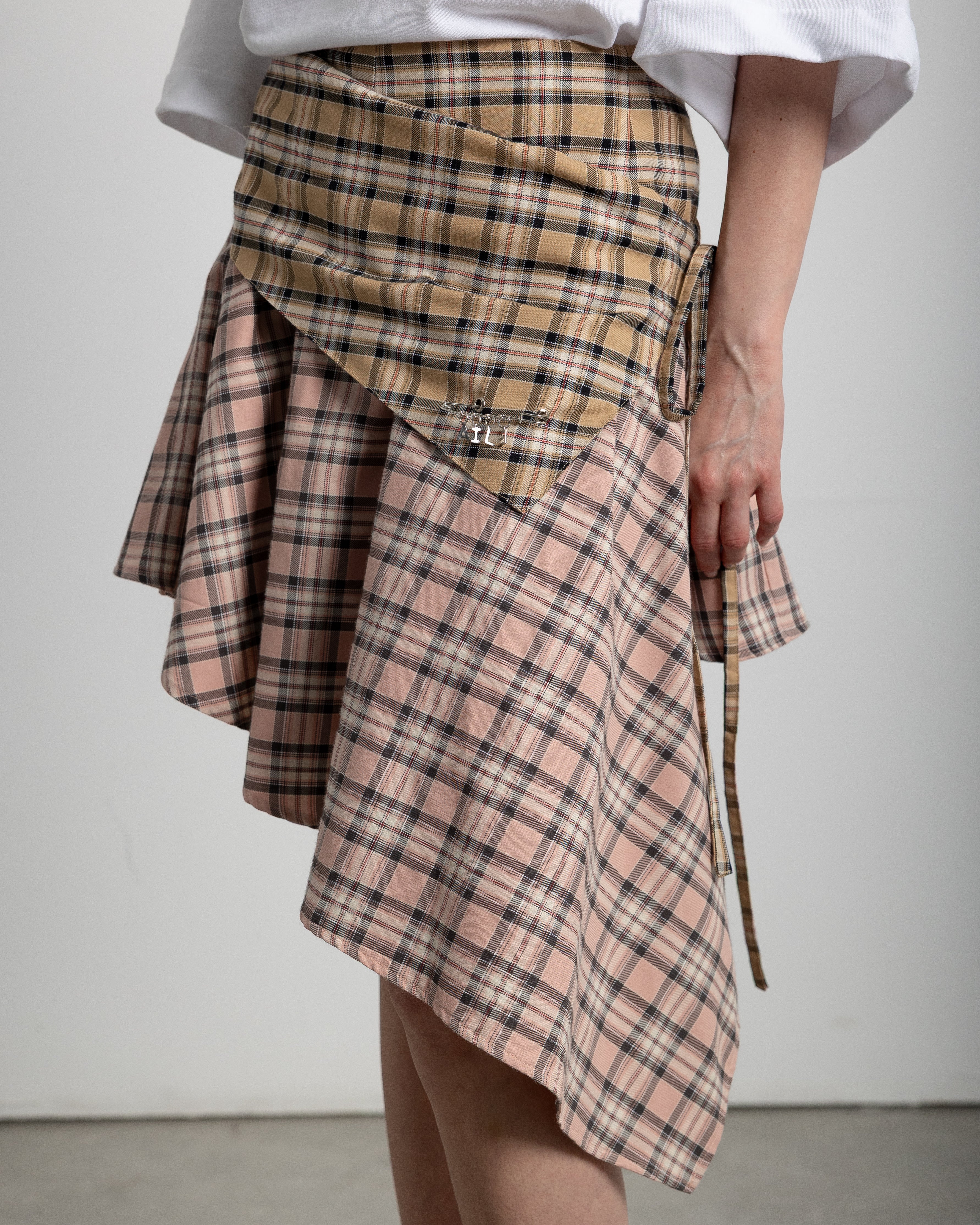 Two-tone asymmetrical tartan skirt. Beige tartan and pink tartan. Fastens with laces on the side. Features a stainless steel pin detail on the front placket