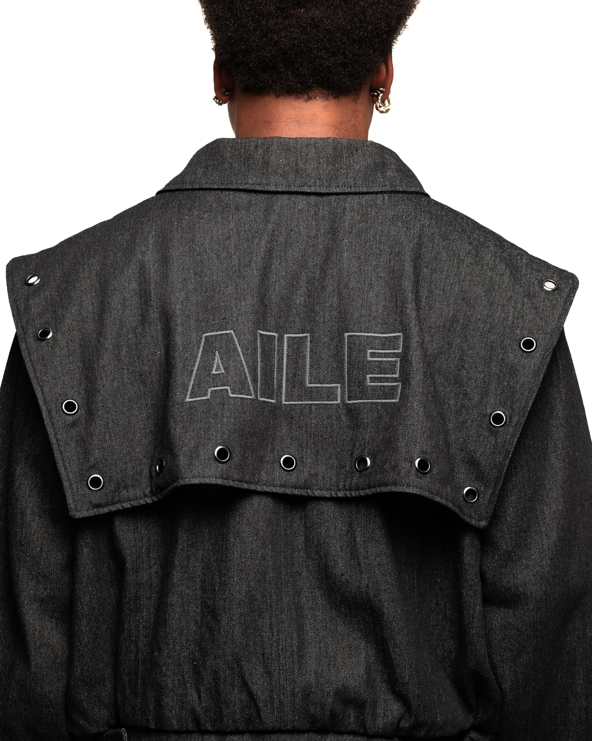 Chaqueta varsity en tejido denim negro con solapa en espalda bordada. Tachuelas a lo largo de toda la solapa, cinturilla con botones ajustables en los costados, puños, cremallera en el delantero y bolsillos delanteros.