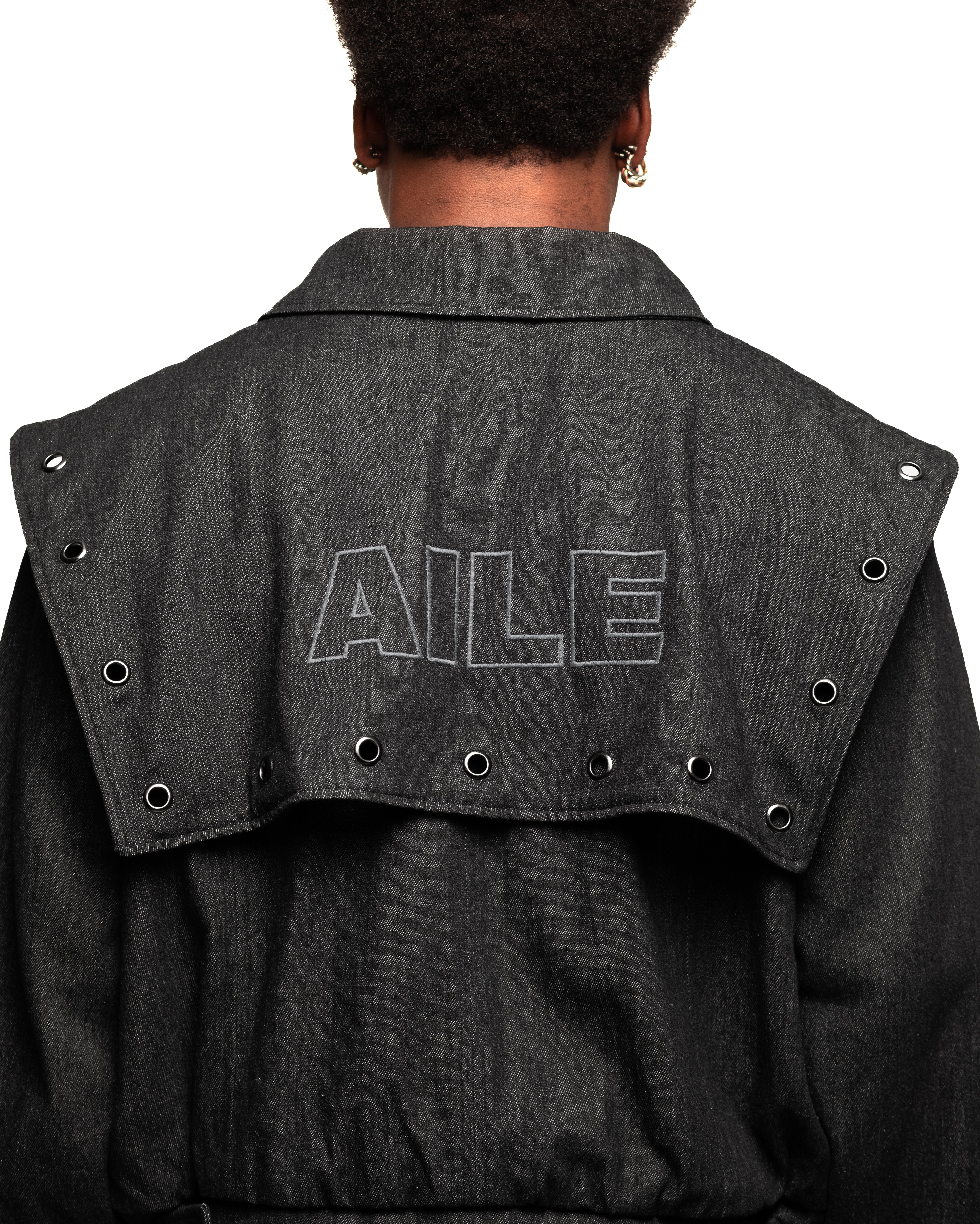 Chaqueta varsity en tejido denim negro con solapa en espalda bordada. Tachuelas a lo largo de toda la solapa, cinturilla con botones ajustables en los costados, puños, cremallera en el delantero y bolsillos delanteros.